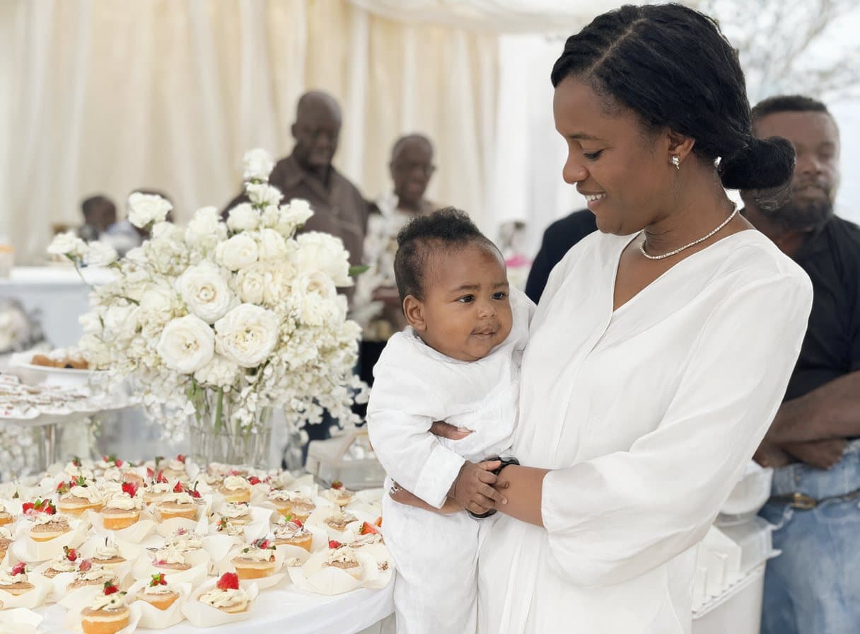 Une maman et son bébé en blanc devant la table des desserts d’un baptême